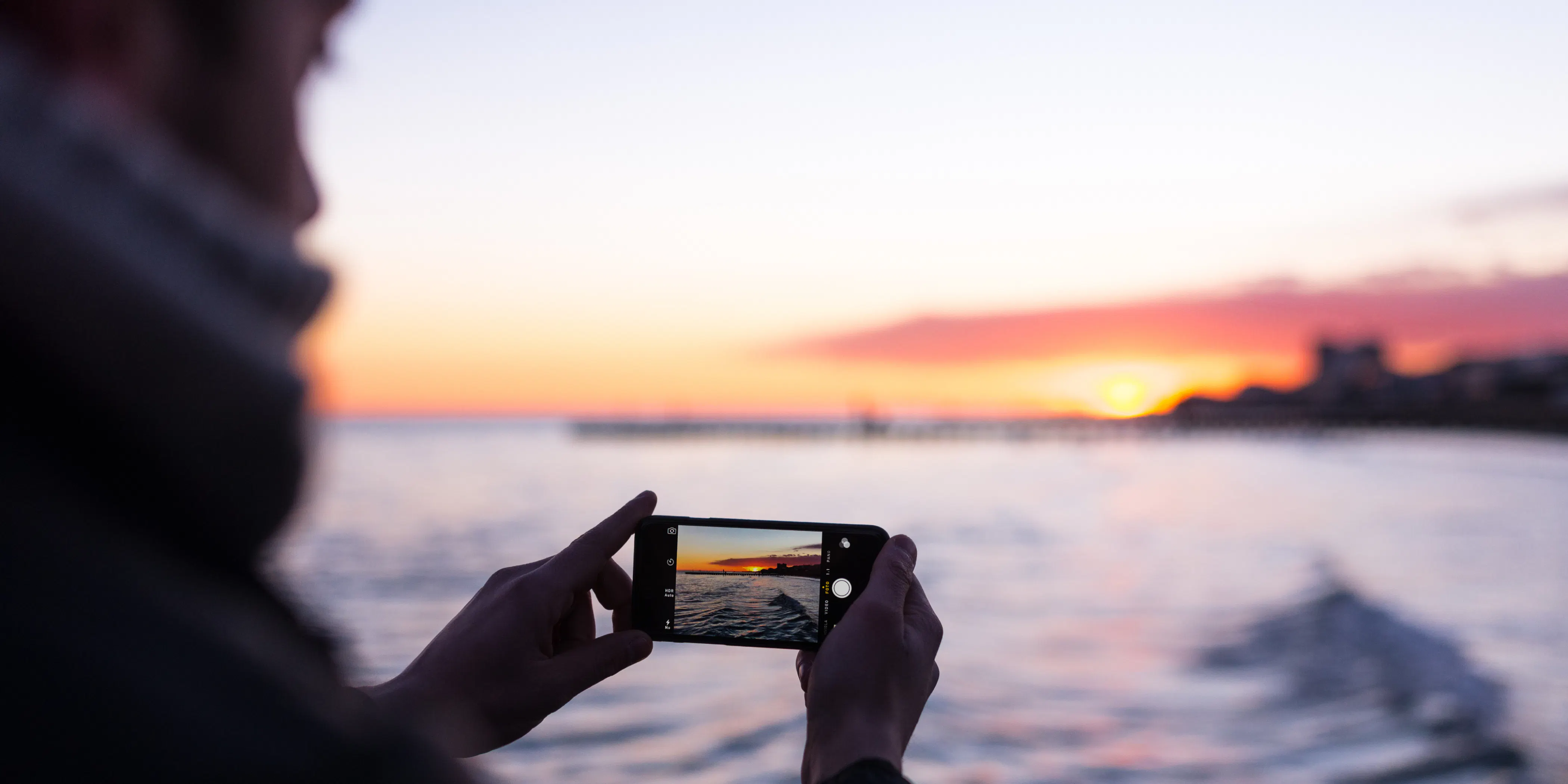 夕暮れの海でスマートフォンを構えて夕日を撮影する人